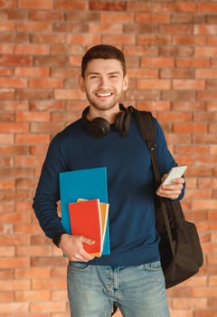 Happy Student Guy With Backpack Holding Cellphone And Copybooks Standing Over Brick Wall Indoor. College Students Lifestyle photo