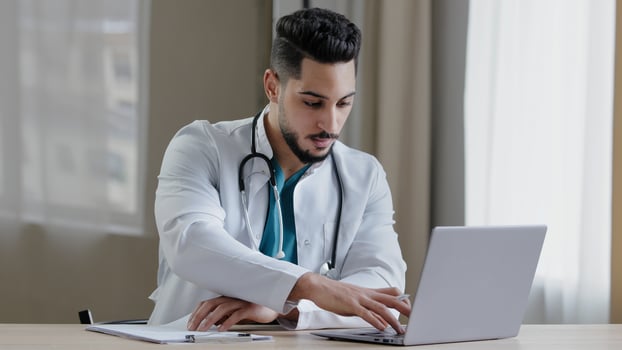 Concentrated arabian man doctor practitioner sit at clinic cabinet use computer chatting distant in medical social app give online consultation writing recipe prescription diagnose search information photo