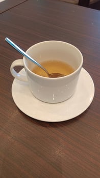A white ceramic cup with a teaspoon in it, filled with light brown tea, rests on a white saucer on a brown wooden table. photo
