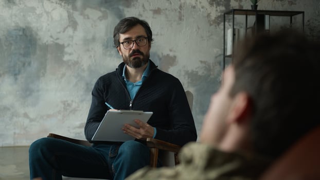Worried mature Caucasian psychologist counselor writing notes listening patient client male officer combat uniform man military army soldier guy veteran lying sofa tell problems at office therapy room photo