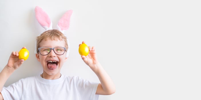Happy child smiles in glasses in a white t-shirt in rabbit ears and with colorful yellow Easter eggs on a white background. Happy Easter concept. photo