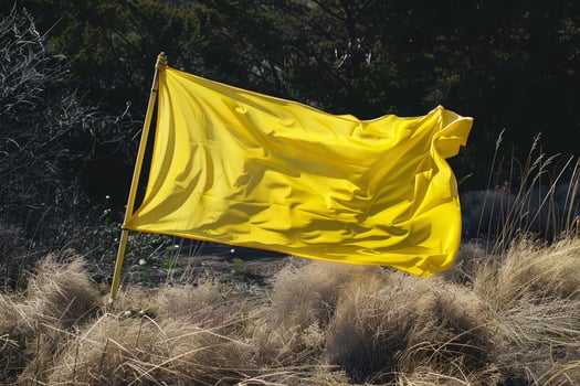 A bright yellow flag stands tall, fluttering in the wind amidst dry grass and scattered greenery, capturing the essence of a sunny day outdoors photo