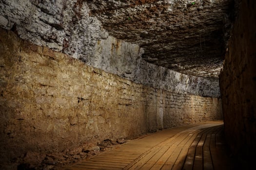 This image depicts a long underground stone tunnel featuring a wooden walkway on the right and rough stone walls illuminated by dim lights, showcasing medieval architecture. photo