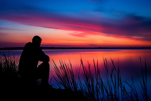 A man sits on a rock by a lake, looking out at the water photo