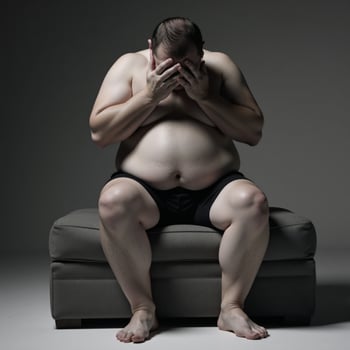 Obese man seated on gray ottoman upset covering face with hands photo