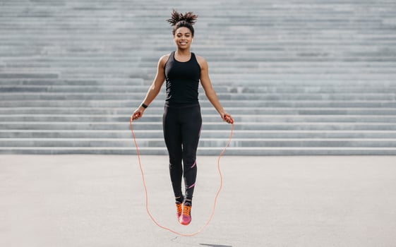 Female athlete does cardio workout. Smiling african american girl in sportswear with fitness tracker jumping rope outdoors, free space photo