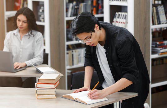 Educational Process. Asian guy writing in notebook, standing at desk in campus library, latin student in the background photo