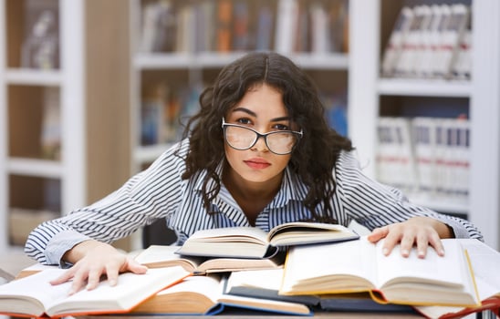 Exhaustion Concept. Frustrated hispanic female student in specs sitting at desk, leaning on books at library, copyspace photo