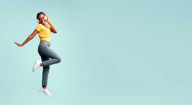 Music App. African Lady In Earphones Jumping Singing Favorite Song Having Fun In Studio Over Blue Background. Free Space photo