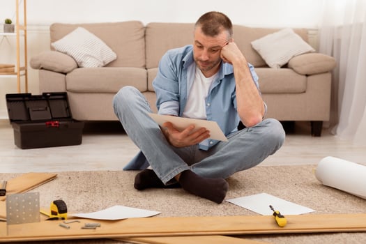 Puzzled Middle-Aged Man Reading Furniture Assembly Manual In Confusion Installing Shelf Sitting Among Self-Assembly Kit Tools And Wooden Parts On Floor At Home. Furniture Assembling Problem photo