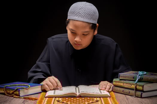 A little Asian Muslim boy is sitting and reading the Quran at the wooden table at his house during the night during the month of Ramadan, Arabic calligraphy meaning of Al Quran. photo