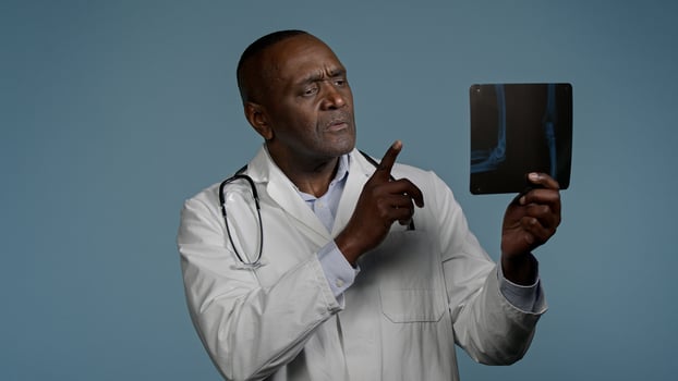 Pensive thoughtful african american man doctor ethnic male radiologist surgeon in studio blue background look at x-ray result checking radiography scan broken bone professional medic analyze trauma photo