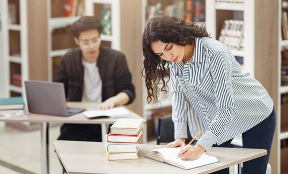 University Life. Mexican teacher writing in notebook, standing at desk in campus library, asian student in the background photo
