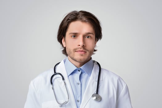 Portrait Of Handsome Young Doctor Wearinf White Medical Coat And Stethoscope And Looking To Camera, Light Studio Background, Copy Space photo