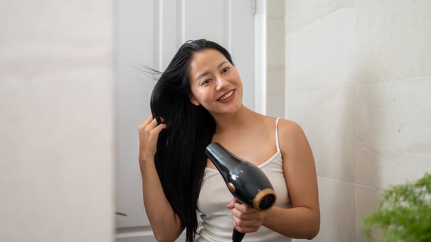 A smiling Asian woman is looking in the mirror, using a hair dryer in the bathroom after shower. photo