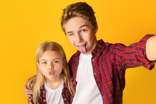 Playful kids taking selfie, little brother and sister making funny faces and fooling together while capturing photo, yellow background, free space photo