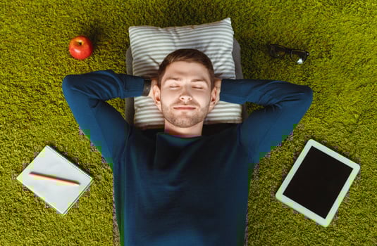 Happy University Student Guy Resting Lying On Green Grass Carpet Among His Belongings Tablet, Notebook. Above View photo