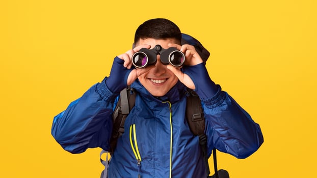 Portrait of smiling young guy hicker using modern binoculars, orange background photo