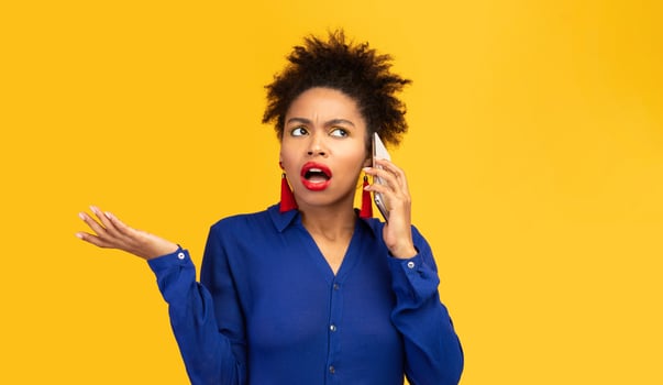 Misunderstanding. Confused black woman talking on mobile phone, girl has many questions isolated on yellow wall photo