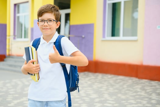 Portrait of little schoolboy in glasses and with backpacks standing on the school building. Study concept photo
