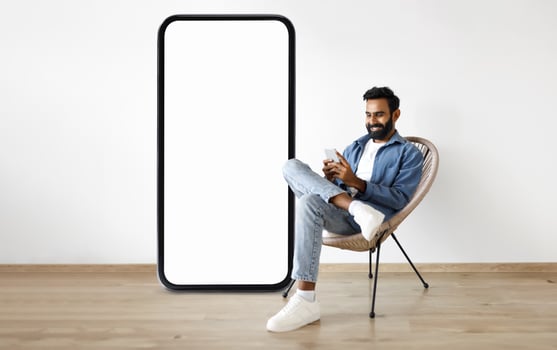 A man sits comfortably in a chair behind large smartphone with blank screen while using his smartphone, surrounded by a modern interior design that emphasizes relaxation and technology. photo