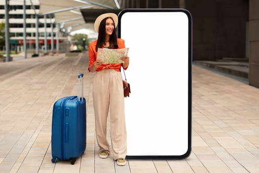 A traveler with a bright outfit holds a map while standing next to a large blank screen, ready to explore the city. A blue suitcase rests beside her in a lively urban area. photo