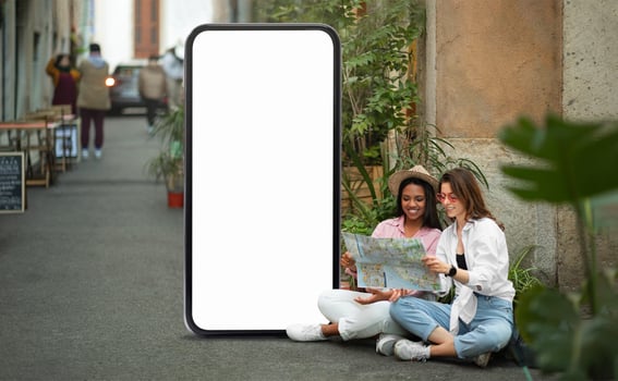Two young women sit together in a lively alley, engrossed in a map. They are surrounded by greenery and a blank phone screen, indicating they are on an urban adventure. photo