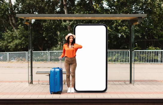 A woman in an orange shirt and hat stands at a bus stop, checking her watch. A large blank screen looms beside her, while her blue suitcase sits nearby, suggesting travel. photo