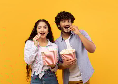 Excited indian man and woman eating popcorn at cinema, standing over yellow studio background. Emotional couple watching comedy movie at cinema or at home, entertainment concept photo