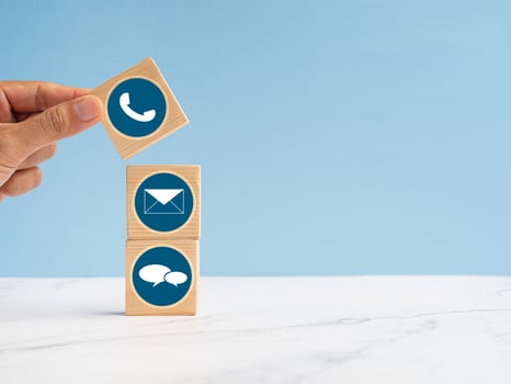 Wooden cubes with symbols of telephone, email, and speech bubble over a marble floor against a light blue background. Contact us concept photo
