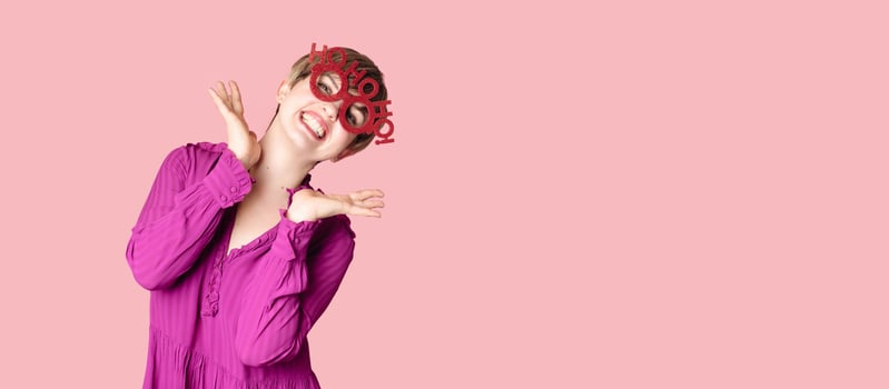 A cheerful young woman in Christmas glasses poses on a highlighted pink background. photo