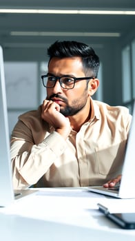 young man with a beard glasses looks pensively at a computer screen photo