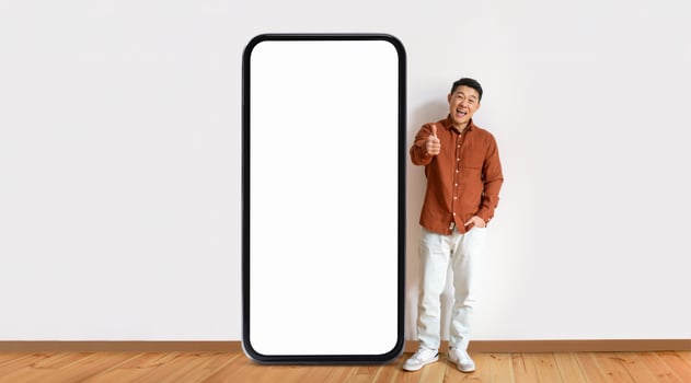 Asian man stands beside a giant blank smartphone, smiling and giving a thumbs up, indicating excitement or approval in a bright and minimalist setting with wooden flooring. photo