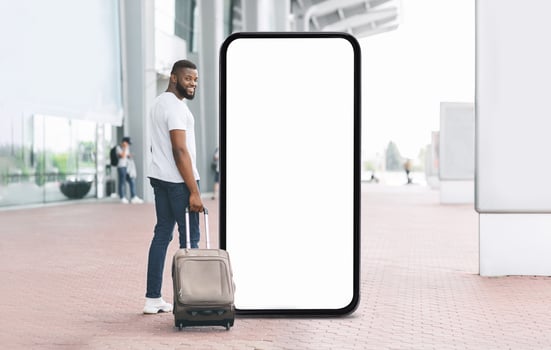 A young black man smiles at the camera while standing beside a large blank screen. He has a suitcase by his side, suggesting he is ready for travel at a busy transportation location. photo