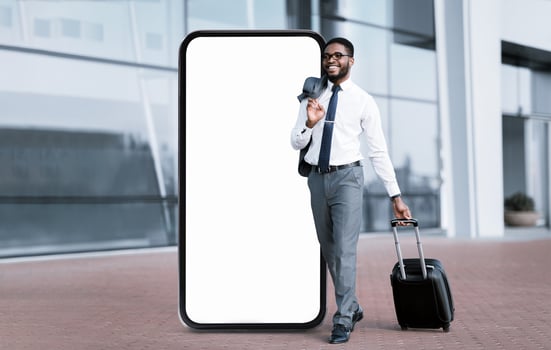 A professional black man in formal wear smiles as he strolls past a giant blank smartphone display, carrying a suitcase. The setting is modern, likely near an office or business center. photo