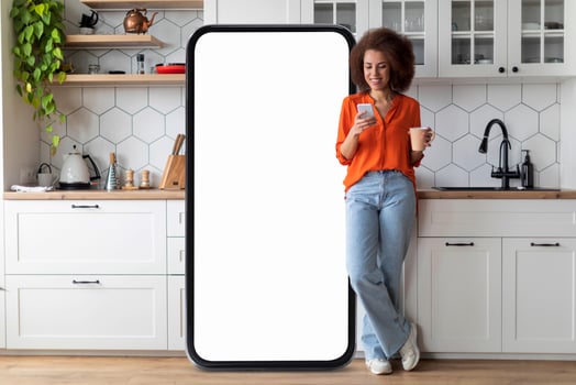 A young black woman in an orange top stands in a chic kitchen, enjoying her coffee and browsing her phone while leaning against a large blank screen. The kitchen features stylish decor. photo