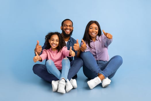 Like Concept. Portrait of smiling black family showing thumbs up gesture with both hands, approving or recommending something good. Mom, dad and daughter sitting on the floor at blue studio photo
