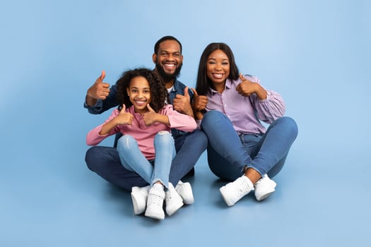 We Approve And Recommend. Excited Black Man, Woman And Teen Girl Gesturing Thumbs Up Okay Looking At Camera Sitting On The Floor Over Blue Background. Studio Shot. Avertisement And Support Concept photo