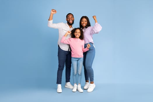 Yes. Full body length of excited young black family of three people shaking clenched fists, making winner gesture. Happy African American man, woman and girl celebrating victory together, blue studio photo