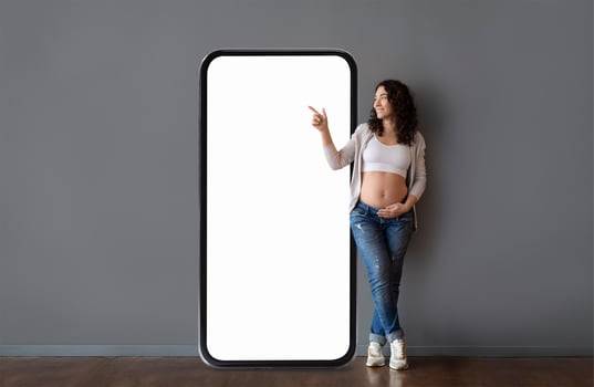 A woman in casual attire stands next to an oversized phone frame, smiling and pointing. She showcases her baby bump, conveying joy and anticipation in a contemporary setting. photo