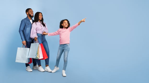 Family Shopping Concept. Full length body size portrait of African American cheerful people holding shopping bags with new clothes, excited little girl pointing aside at copy space, blue studio wall photo