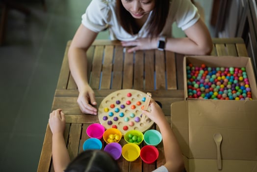 Asian woman and a child are sitting at a wooden table, playing a game with colorful balls. The woman is helping the child with the game, and they are both smiling and enjoying themselves photo