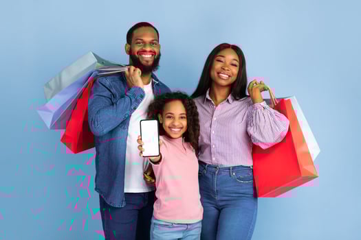 Portrait of happy family of three people of color holding shopping bags, cheerful girl showing smartphone with white empty screen in hand, isolated on blue studio background, mock up, blank space photo