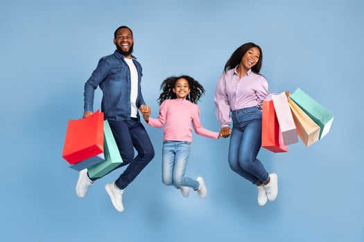 Sales And Discounts. Full body length shot of excited African American family of three people jumping, holding hands and shopping bags with new clothes, rejoice purchase, blue studio wall photo