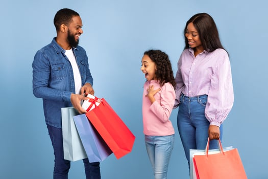 Congratulation. Portrait of smiling African American dad and mum making present for their daughter, man taking out gift box with present from colorful shopping bag for surprised girl, blue studio wall photo