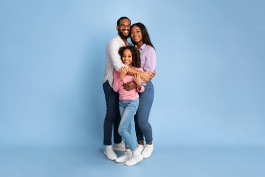 Happy Loving Family. Vertical full body length shot of African American woman and man embracing smiling girl standing isolated on blue studio wall. Cheerful mother and father posing with daughter photo