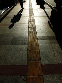 A person is walking on a sidewalk next to a bench. The bench is empty. The sidewalk is lined with bricks and has a shadow cast by the person photo