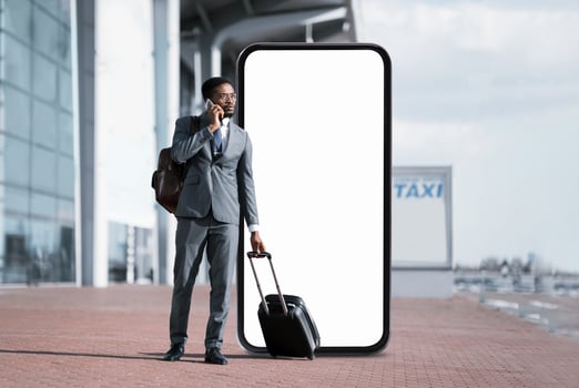 African American man in a suit stands at the airport terminal next to huge phone with blank screen, talking on his smartphone while holding a suitcase. photo