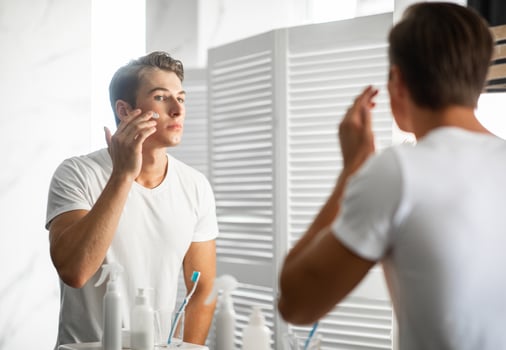Male Facial Skincare Concept. Portrait Of Confident Handsome Man Applying Moisturizing Cream On Smooth Face Caring For Skin After Shaving Routine, Looking In The Mirror, Standing In Bathroom At Home photo