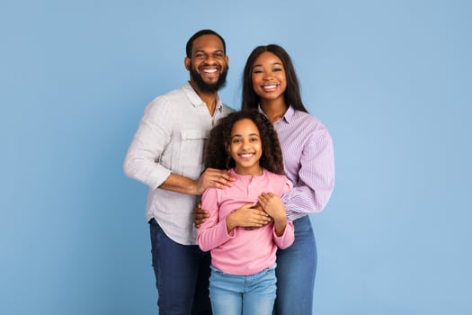 Love And Care Concept. Portrait of African American mum and dad embracing smiling daughter standing isolated on blue studio background. Cheerful man and woman posing with girl and looking at camera photo